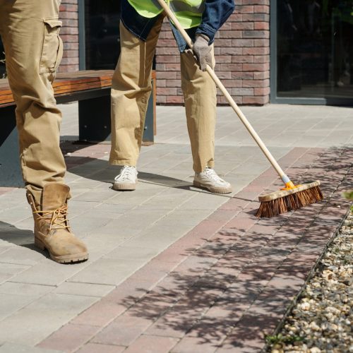 Two middle aged men cleaning outdoor walkway, one holding trash bag while other sweeping pavement with broom, both wearing work boots and gloves, performing maintenance near building